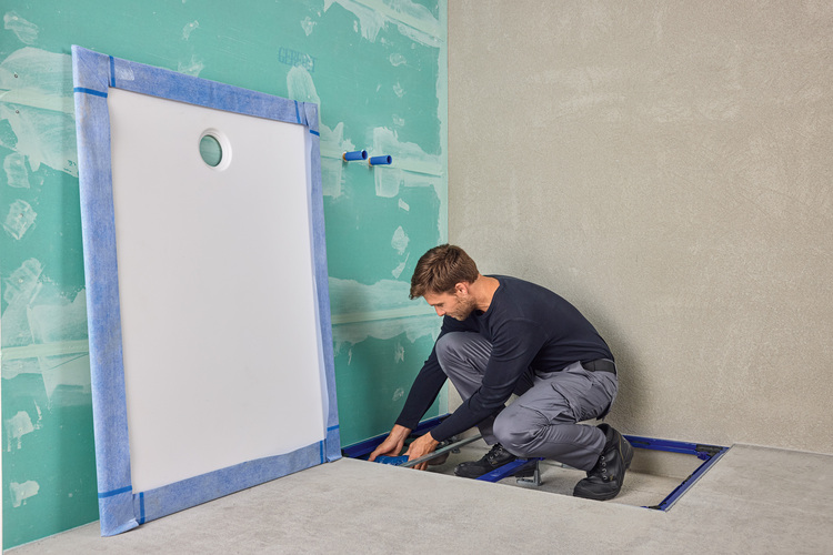 Fachhandwerker montiert bodengleiche Duschwanne auf Rohbauboden, mit Werkzeug; unbeplankte Leichtbauwände kennzeichnen den Baufortschritt.