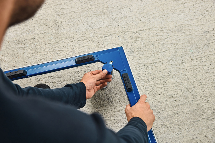 Montage eines blauen Metallrahmens für Gebäudetechnik; Fachkraft befestigt Eckstück auf Betonboden, typische Arbeit im SHK-Bereich.