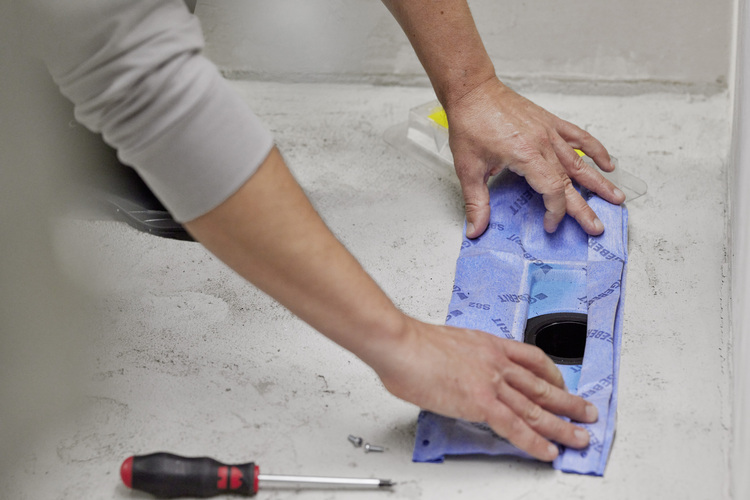 Fachkraft bringt blaues Abdichtungsbahn-System fachgerecht um Bodenablauf auf Betonboden an; typische Werkzeuge für SHK-Montage liegen bereit.