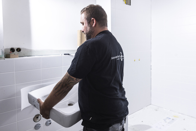 Fachhandwerker montiert ein weißes Waschbecken an einer gefliesten Badezimmerwand - Beispiel für Sanitärinstallation in der Gebäudetechnik.