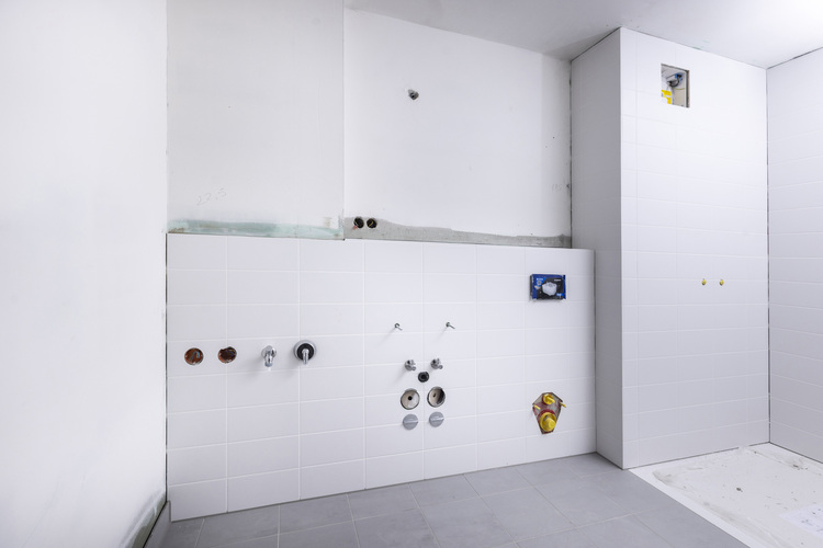 Sanitärrohinstallation: Teilweise gefliestes Bad während Bauphase, sichtbare Rohrleitungen und Armaturen vor vorbereiteter weißer Installationswand.