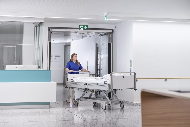 Modernes Krankenhaus: Pflegekraft bewegt leeres Patientenbett durch lichtdurchfluteten, hygienischen Flur - Fokus auf Gebäudetechnik und Facility Management.