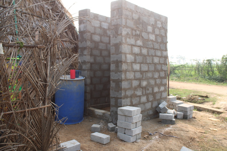 Unfertiger Sanitärraum aus Ziegeln neben einem Strohdachbau, sichtbarem Wasserfass und Baumaterial - Beispiel für Rohbau in der Gebäudeinstallation.