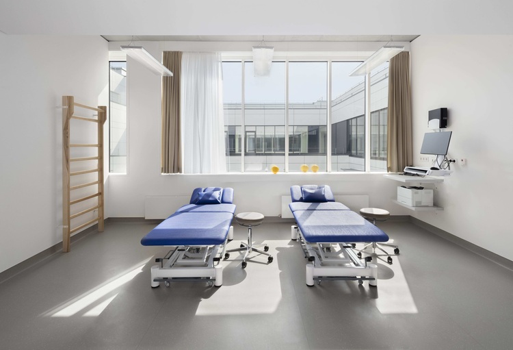 Lichtdurchfluteter Therapieraum mit zwei blauen Behandlungsliegen und großen Fenstern, Beispiel für barrierefreie Raumgestaltung in Gesundheitsbauten.