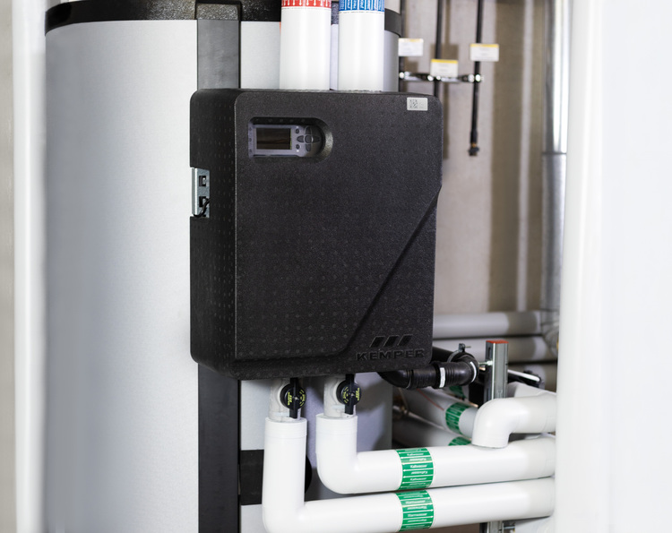 Schwarze Regelungseinheit mit angeschlossenen Rohrleitungen in einem Technikraum, installiert neben einem großen zylindrischen Warmwasserspeicher.