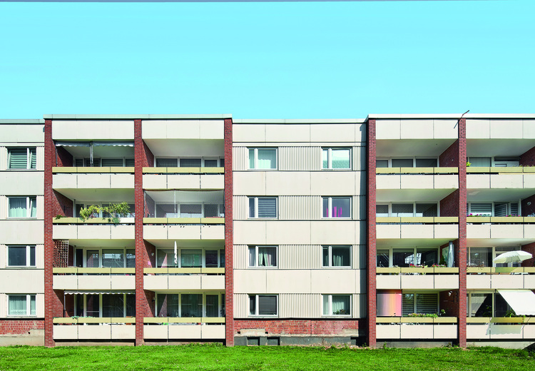 Modernes viergeschossiges Wohngebäude mit Balkonen, großen Fenstern und energieeffizienter Gebäudetechnik, vor blauem Himmel und Rasenfläche.