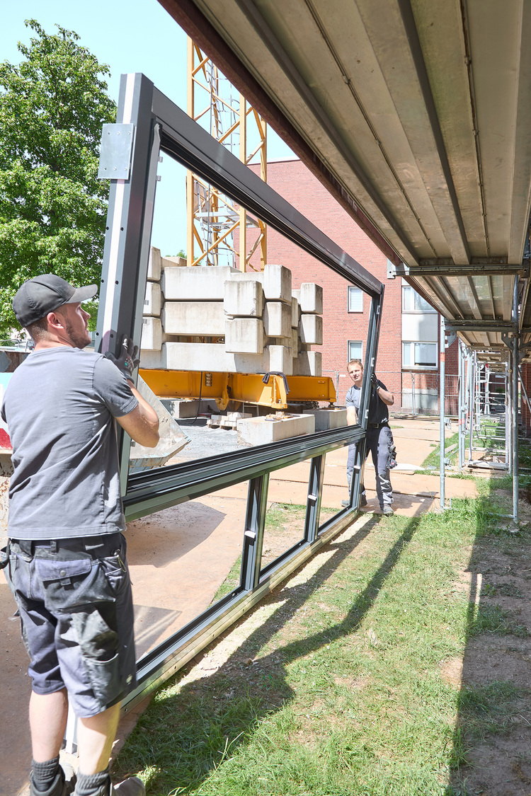 Zwei Monteure transportieren einen großen Metall-Fensterrahmen auf einer Baustelle mit Gerüst - Beispiel für moderne Gebäudehüllentechnik.