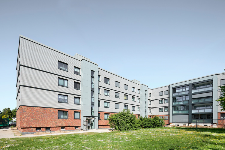 Viergeschossiges Mehrfamilienhaus mit moderner Fassade aus grauen Paneelen und Klinker, umgeben von begrünten Außenflächen - Beispiel energieeffizienter Neubau.