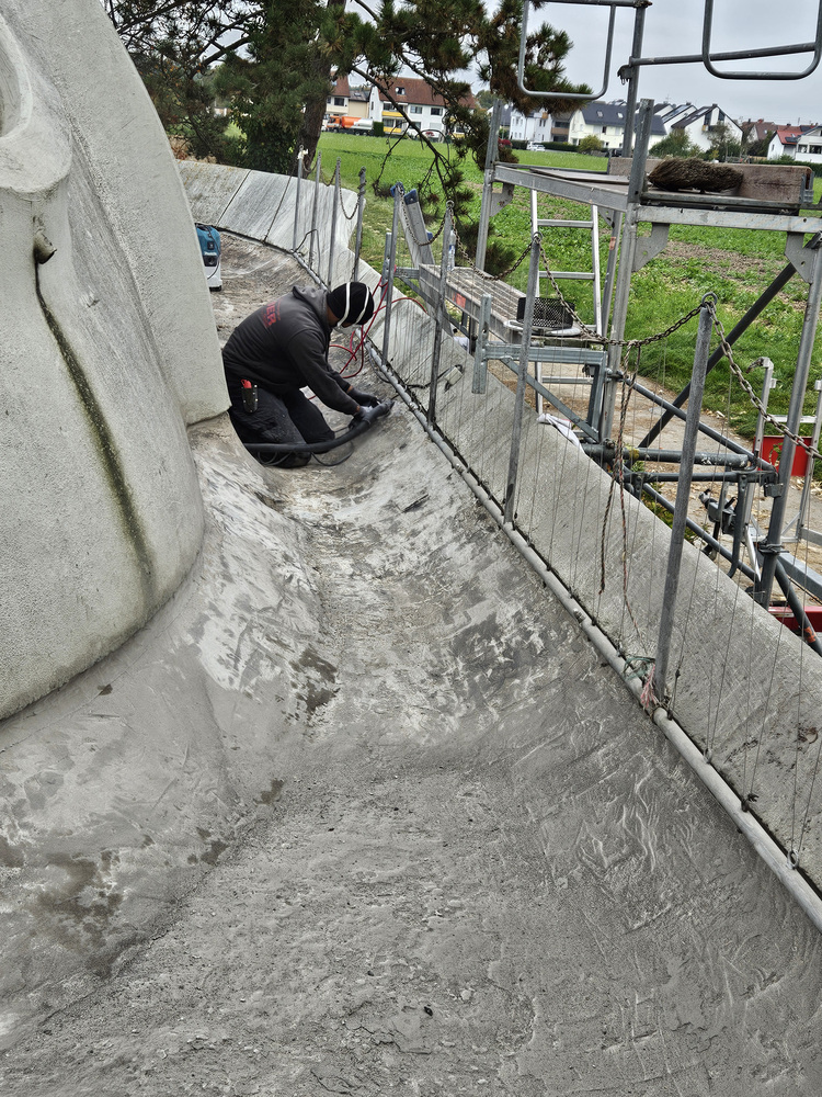 Facharbeiter mit PSA bei Instandsetzung einer geschwungenen Betongebäudestruktur, umgeben von Baugerüst und Absperrungen auf Baustelle der Gebäudetechnik.