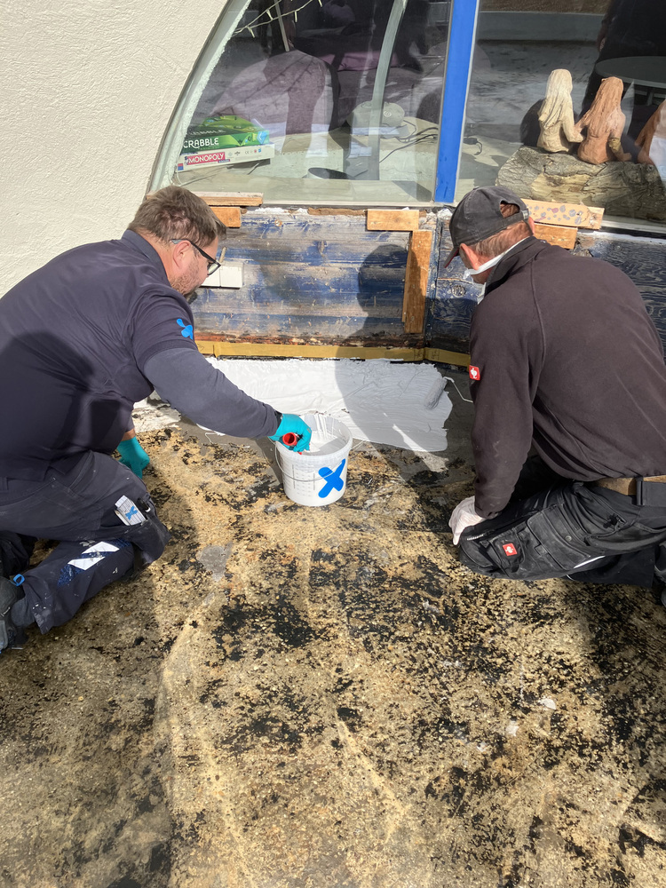 Fachkräfte tragen weißen Dichtstoff mit Pinsel an einer Fensterlaibung auf - Abdichtung und energetische Modernisierung in der Gebäudetechnik.