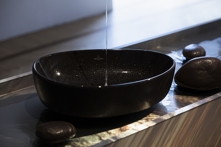 Modernes schwarzes, schalenförmiges Waschbecken mit Wasserzufluss und schwarzen Ziersteinen, beispielhaft für zeitgemäße Sanitärtechnik im Objektbau.