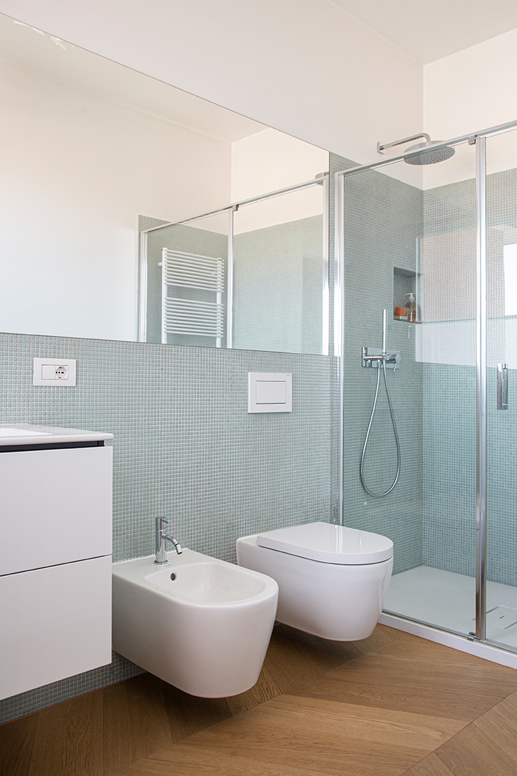 Barrierearmes Bad mit wandhängendem WC, Bidet, bodengleicher Dusche, weißem Waschtisch und hellem Holz-Heringsboneboden für moderne Gebäudetechnik.