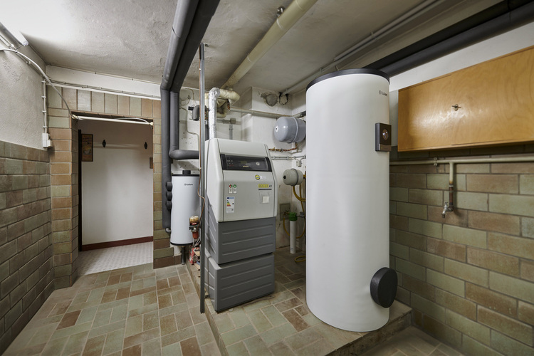 Technikraum im Keller mit Warmwasserspeicher, Heizkessel und Rohrleitungen; gefliester Boden und Wände, typische Installationstechnik.