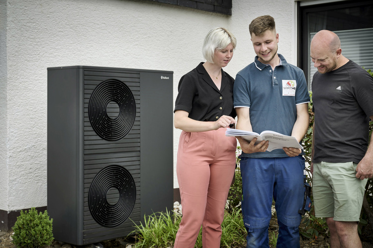 Drei Fachkräfte besprechen technische Unterlagen an einer außen installierten Wärmepumpe vor einem Wohngebäude, Fokus auf Gebäudetechnik.