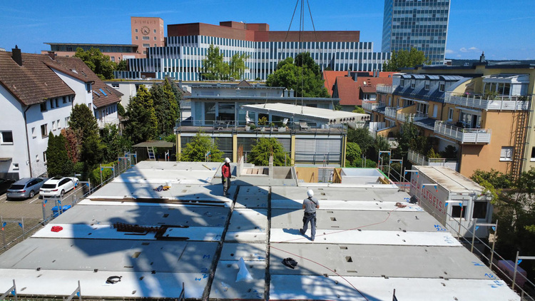 Zwei Bauarbeiter mit Schutzhelmen auf einer Gebäudetechnik-Baustelle, im Hintergrund städtische Gebäude und Wohnhäuser.