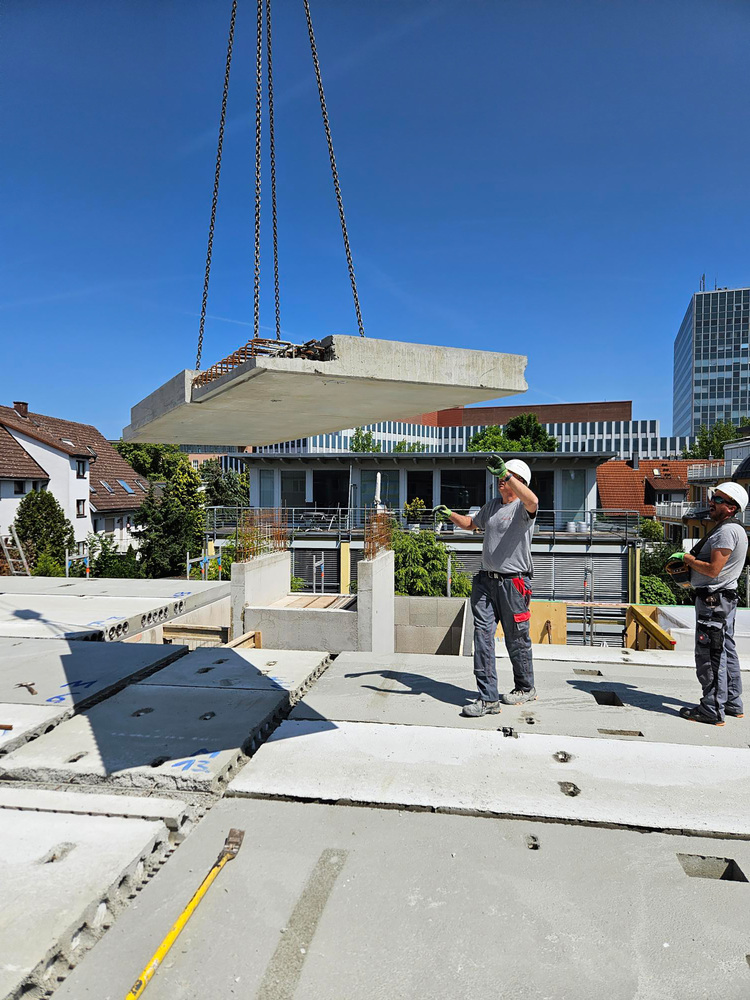 Zwei Facharbeiter positionieren eine Betondecke, die auf einer TGA-Baustelle pro Kran eingehoben wird, für den Hochbau im Gebäudetechniksektor.
