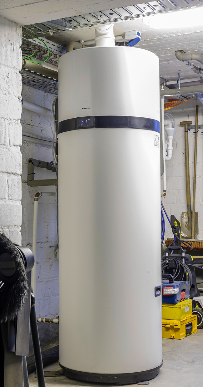 Stehender weißer Warmwasserspeicher mit angeschlossenen Rohrleitungen und Werkzeugen in einem Technikraum, typische Installation im Gebäudetechnikbereich.