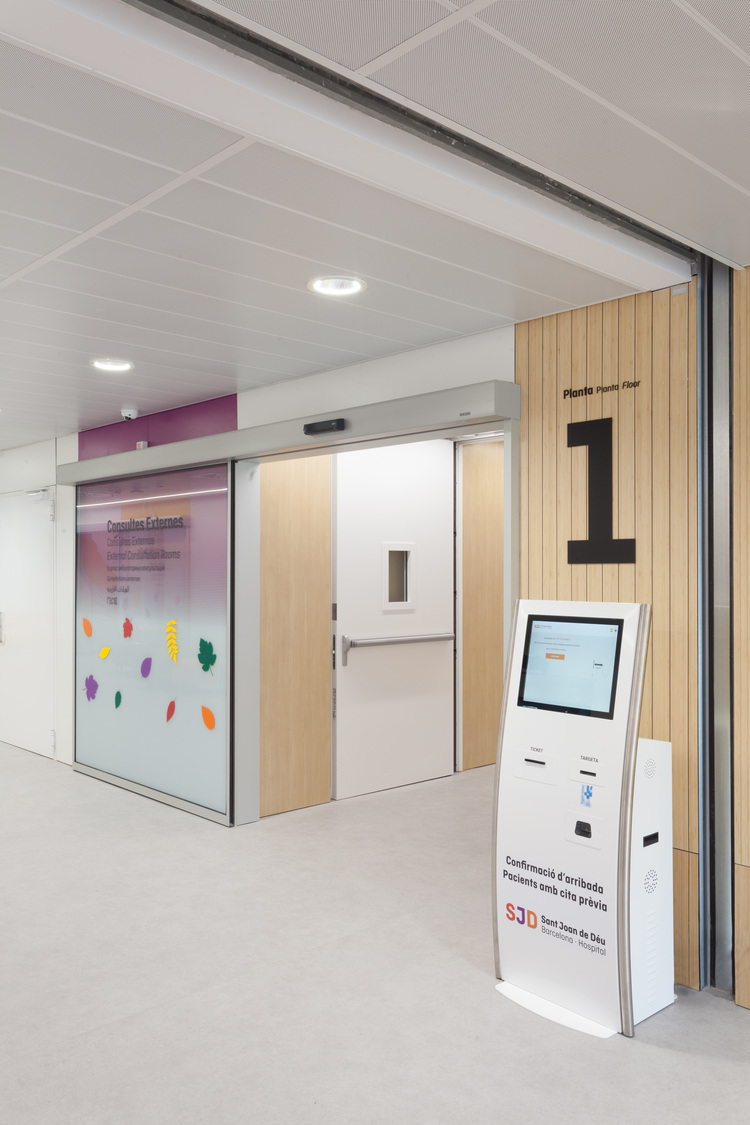 Barrierefreie Krankenhausflur mit digitalem Self-Service-Check-in-Terminal, großer Zonenkennzeichnung "1" und gläserner Wand mit Blätterdekor.