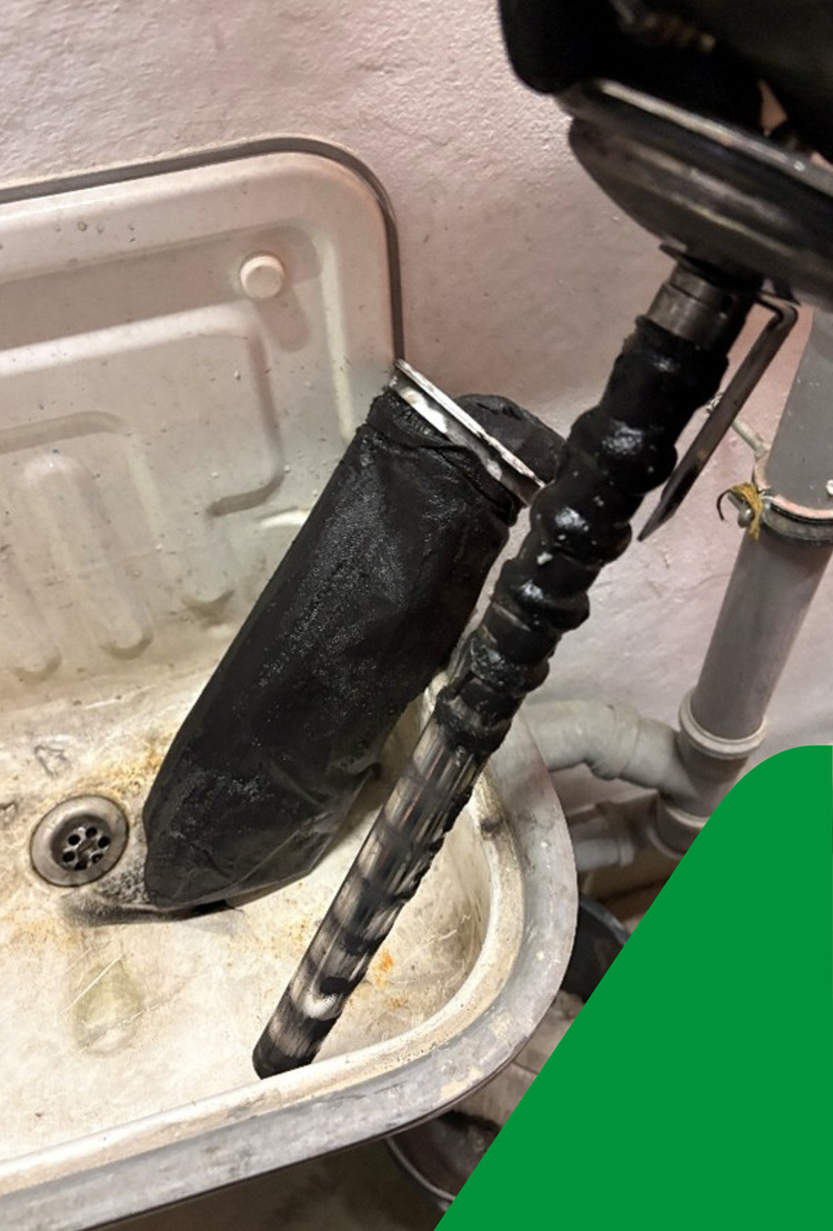 Ein verschmutzter schwarzer Filter und eine Metallstange liegen im Ausgussbecken; sichtbare Rohrleitungen und Ablauf, typisch für Wartungsarbeiten.