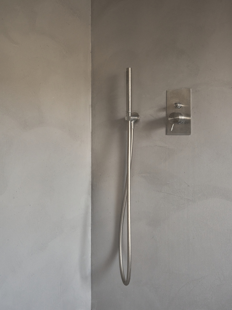 Moderne Duschinstallation mit Edelstahl-Handbrause und Steuerungspanel an glatter grauer Wand, geeignet für barrierefreie Sanitärkonzepte im Objektbau.