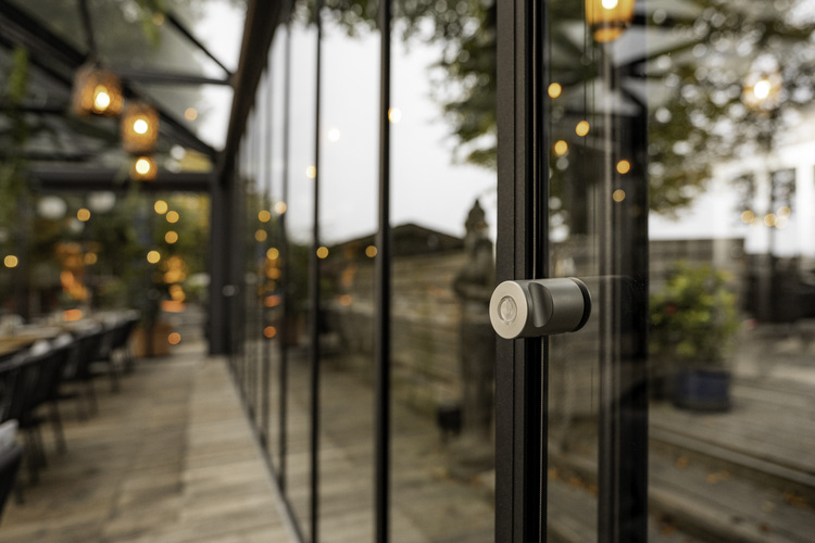 Verglaste Tür mit rundem Edelstahlgriff, spiegelt Außenbeleuchtung; dahinter Terrasse mit Sitzbereich, geeignet für moderne Gebäudezugänge.