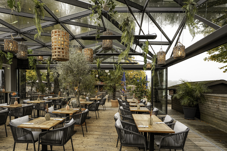 Modernes Restaurant mit Glasdach, Holztischen und Hängepflanzen, zeigt Beispiel für nachhaltige Architektur und moderne Gebäudetechnik in der Gastronomie.