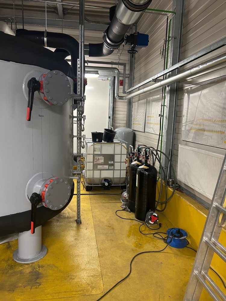 Technikraum einer Gebäudetechnik-Anlage mit groß dimensionierten Pufferspeichern, Rohrleitungen, Armaturen und Steuerungstechnik auf gelbem Boden.
