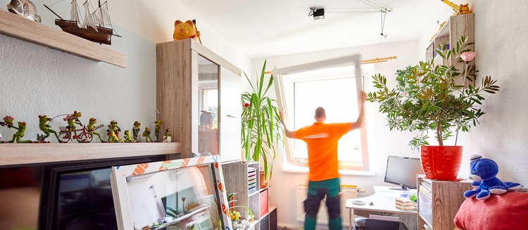 Fachkraft im orangefarbenen Shirt montiert Fenster in hellem Raum, relevante Tätigkeit für Gebäudetechnik und energieeffiziente Gebäudehülle.