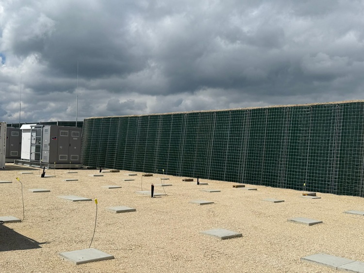 Außenbereich einer Gebäudetechnik-Anlage mit Schotterbelag, Betonfundamenten, Rohrleitungen und großem Schutzgitter; die relevante Infrastruktur ist sichtbar.