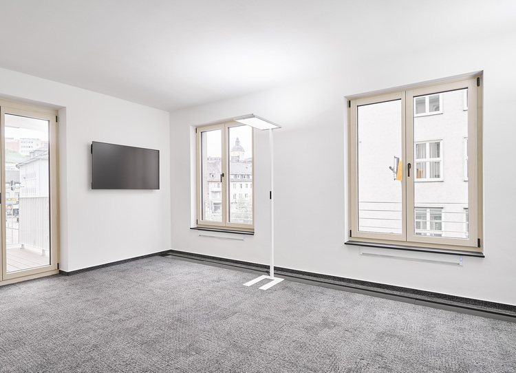 Moderner, minimalistisch eingerichteter Innenraum mit grauem Teppichboden, zwei Fenstern mit Ausblick und Wand-TV, Beispiel für energieeffiziente Gebäudetechnik.