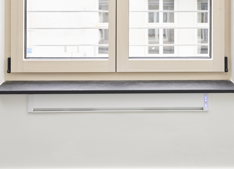 Weißes Wohnraum-Lüftungsgerät unter geschlossenem Fenster mit beigem Rahmen und schwarzer Fensterbank, installiert in moderner Gebäudehülle.