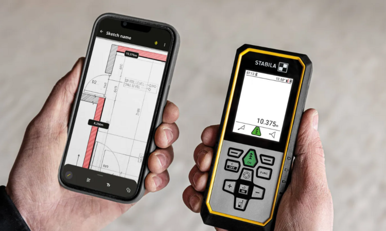 Ein Fachhandwerker vermisst einen Raum präzise mit einem digitalen Entfernungsmesser; auf dem Smartphone wird der Grundriss des Gebäudes angezeigt, um die Daten exakt zu erfassen.