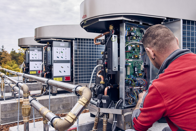 Ein SHK-Fachmann wartet eine geöffnete Split-Klimaanlage im Außenbereich, wobei die inneren Bauteile sichtbar sind; im Hintergrund sind zwei Klimageräte zu sehen.