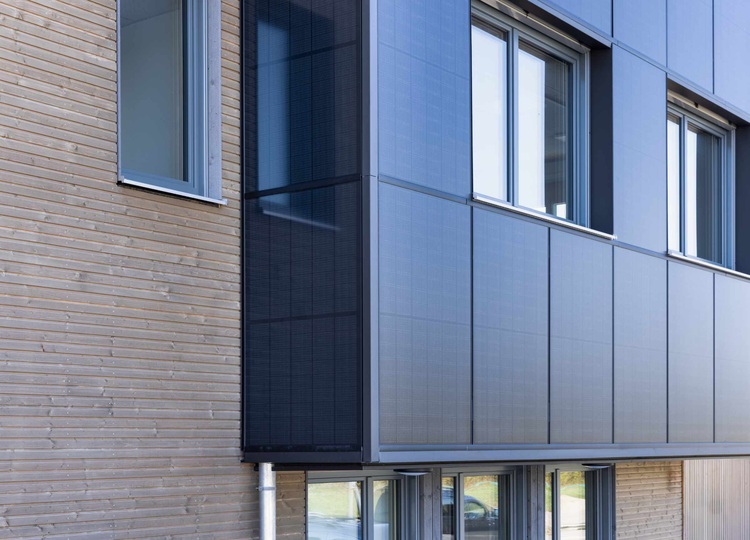 Fassade eines modernen Gebäudes mit vertikalen Holzlamellen auf der linken Seite und PV-Modulen auf der rechten Seite, große Fenster; Beispiel für eine energieeffiziente Gebäudehülle.