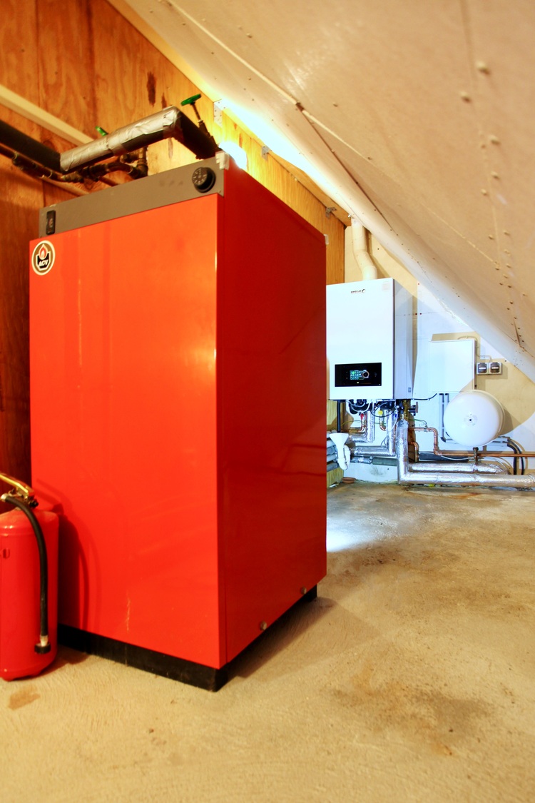 Rote Heizkesselanlage mit Feuerlöscher, offenen Rohrleitungen und weißem Steuerungsmodul in einem Technikraum – eine typische gebäudetechnische Anlage.