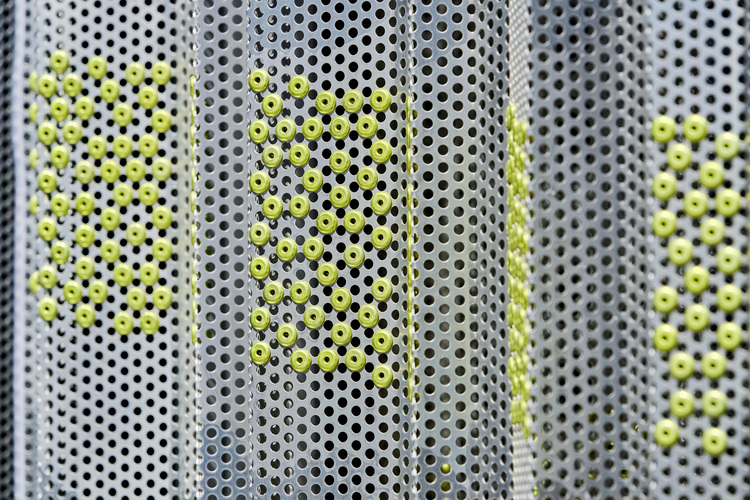 Detailansicht einer perforierten Metallfassade mit gelben, runden Elementen in rechteckigen Mustern – ein Beispiel für eine innovative Gebäudehülle im Bereich der Gebäudetechnik.
