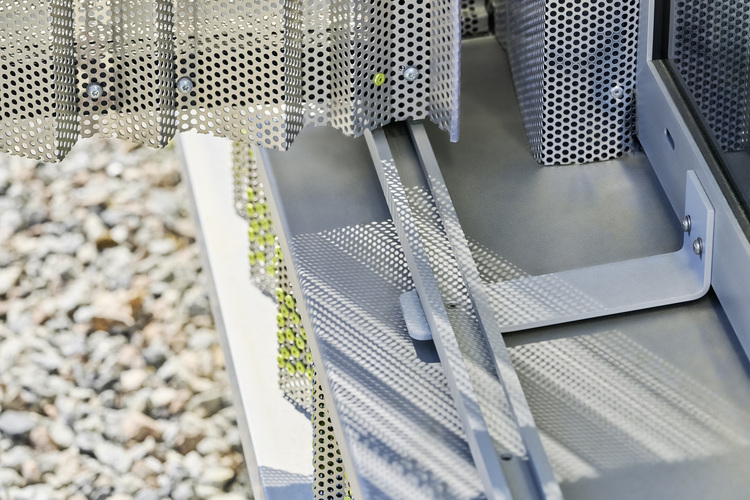 Perforierte Metallpaneele als Fassadenelemente werfen Schatten auf die graue Bodenfläche; im Hintergrund Kies – ein Beispiel für moderne Gebäudehüllen.