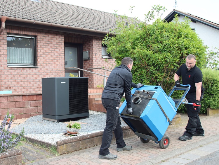 Zwei Fachkräfte transportieren ein großes blaues HLK-Gerät auf einem Rollwagen vor einem Backsteinhaus, in dessen Garten eine Wärmepumpe steht.