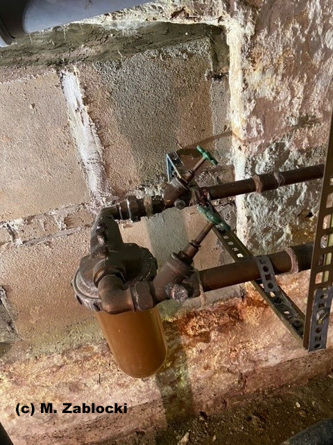 Korrodierter Wasserfilter und Metallrohre an einer Betonwand in einem Technikraum; Beispiel für mangelhafte Wartung in der Gebäudetechnik.