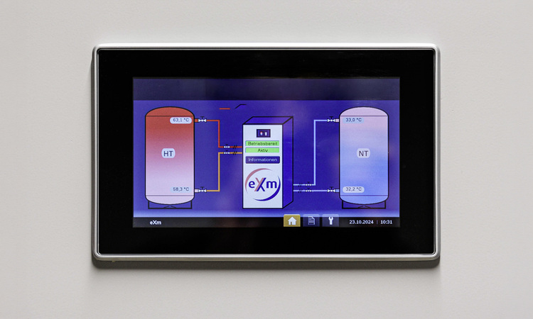 Touchpanel einer Gebäudeleittechnik, das auf einer blauen Benutzeroberfläche eine Anlagenübersicht mit Tanks, Temperaturanzeigen und Systemdaten anzeigt.