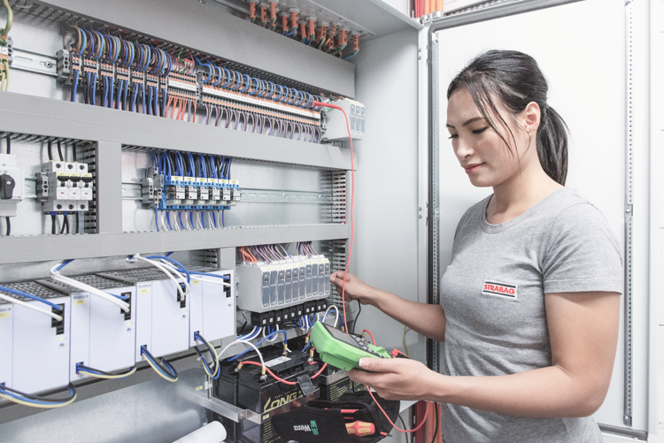 Ein Fachmann prüft mit einem Multimeter ein komplexes Schaltschrankmodul in der Gebäudetechnik; zahlreiche farbige Kabel sind zu sehen.