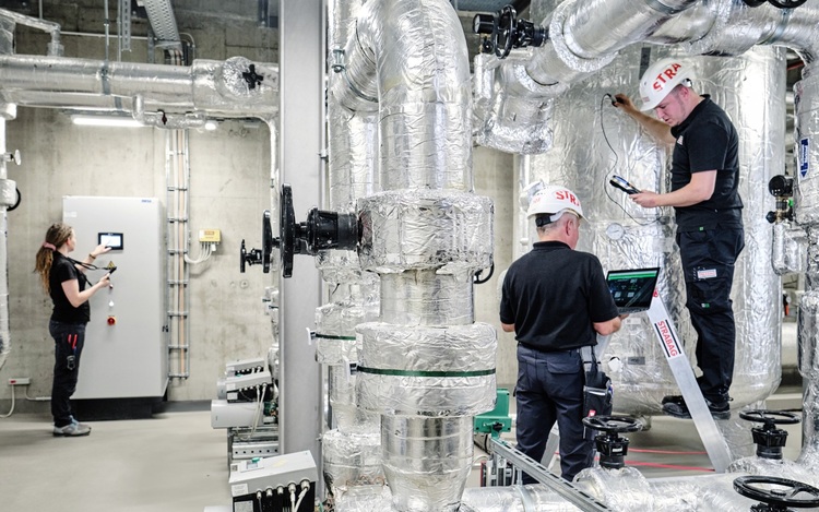 Drei Fachkräfte überprüfen und warten große, isolierte Rohrleitungen in einer haustechnischen Anlage für Heizung oder Klimatisierung.