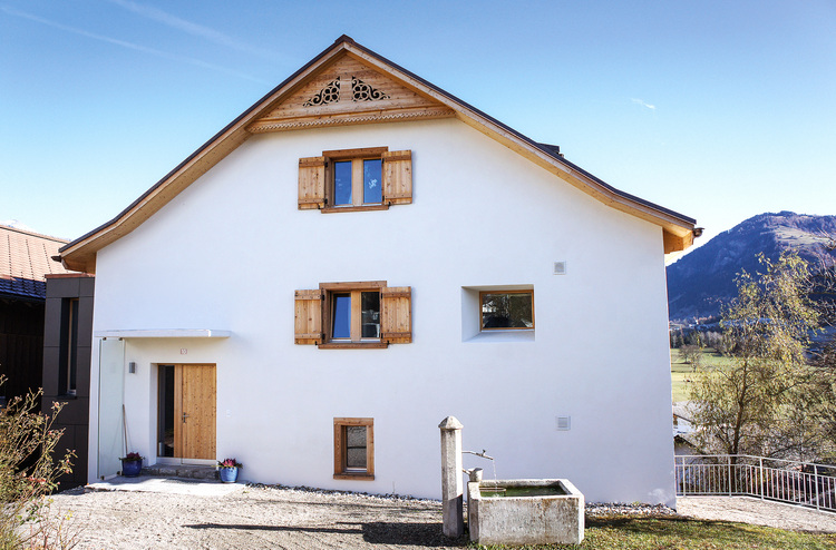 Modernes Einfamilienhaus mit gedämmter Gebäudehülle, Holzfensterläden und Satteldach, Vorgarten mit Natursteinbrunnen, Berge im Hintergrund.