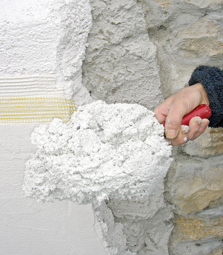 Ein Facharbeiter trägt mit einer Kelle eine weiße Putzschicht auf eine unebene Steinwand auf – ein Arbeitsschritt bei der Gebäudesanierung zur Steigerung der Energieeffizienz.