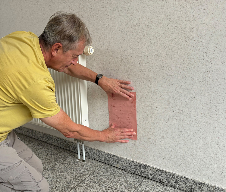 Ein Facharbeiter bringt eine Wanddämmplatte auf Keramikbasis im Rahmen einer energetischen Sanierung an; weiß gestrichene Wand, Heizkörper im Hintergrund.