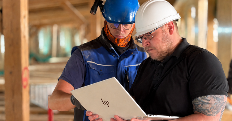 Zwei Fachkräfte mit Schutzhelmen prüfen auf der Baustelle eines Gebäudetechnik-Projekts im Rohbau Pläne am Laptop.