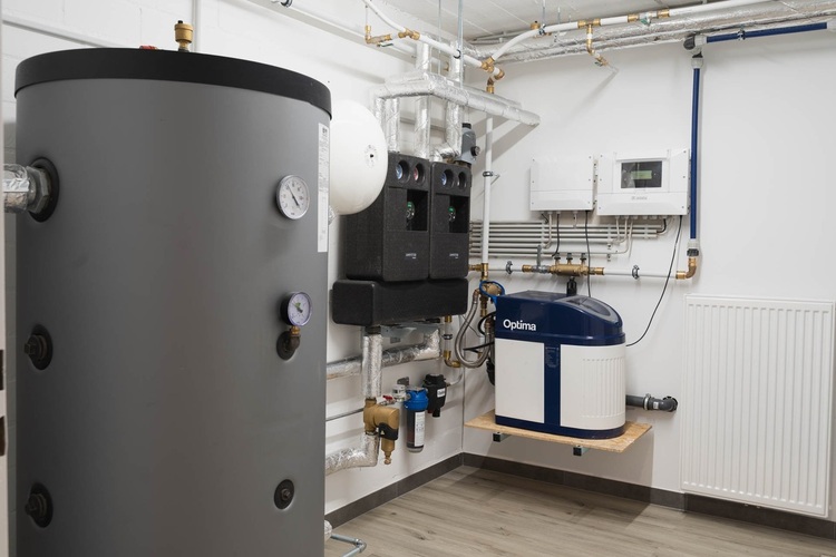 Heizungsraum mit großem Warmwasserspeicher, Steuerung, Messgeräten, Rohrleitungen und Optima-Einheit auf hellem Holzboden.