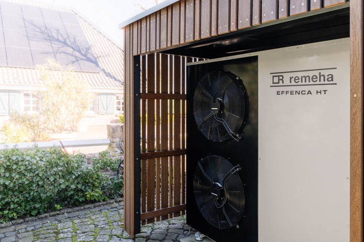 Luft-Wärmepumpen-Außengerät mit zwei Ventilatoren unter einem Holzvordach, neben dem Garten und dem Gebäude mit PV-Anlage – energieeffiziente Gebäudetechnik.