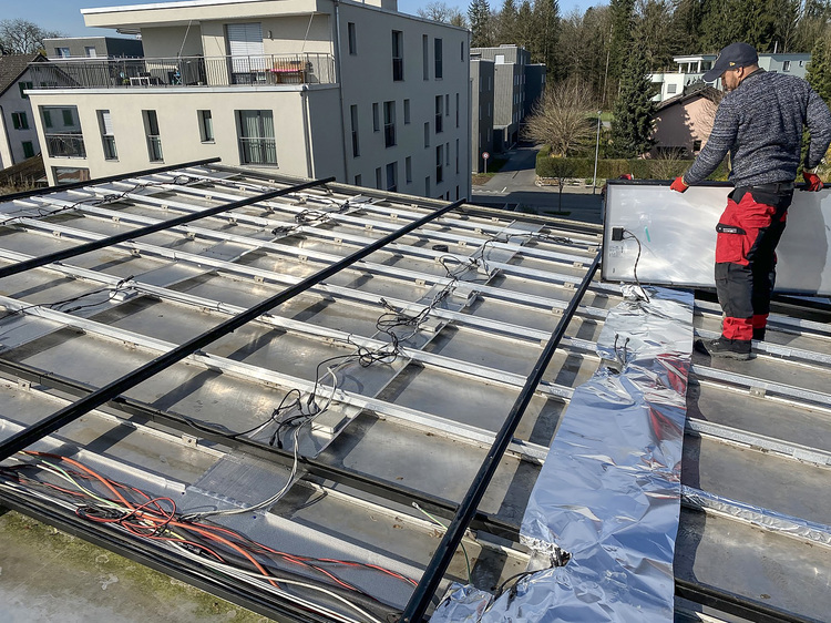Ein Fachmann montiert Photovoltaikmodule auf einer Dachkonstruktion, die Verkabelung ist sichtbar; im Hintergrund sind moderne Wohngebäude zu sehen, der Fokus liegt auf der Gebäudetechnik.