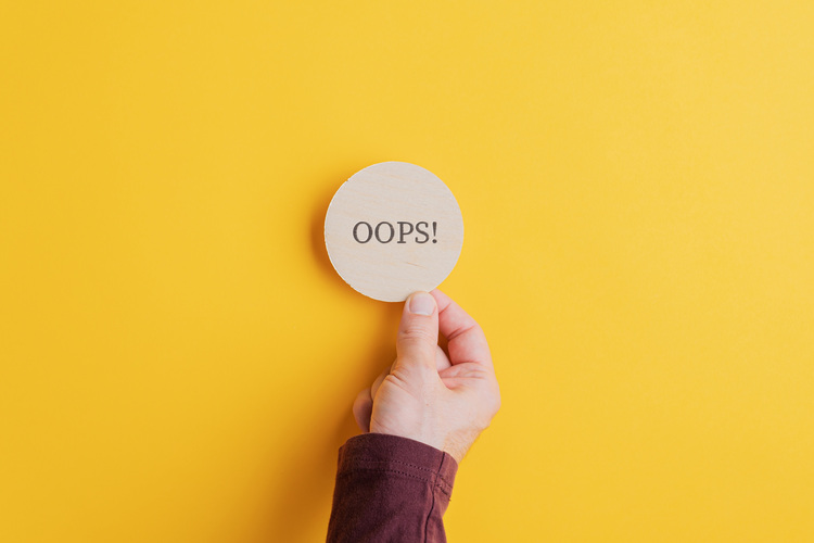 Eine Hand hält ein rundes Holzschild mit der Aufschrift „OOPS!“ vor einem gelben Hintergrund – ein Symbol für Fehler oder Störungen in der Gebäudetechnik.